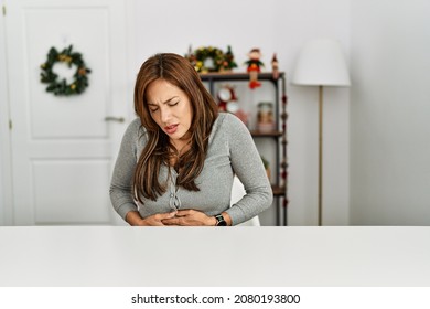 Young Latin Woman Sitting On The Table By Christmas Decor With Hand On Stomach Because Indigestion, Painful Illness Feeling Unwell. Ache Concept. 