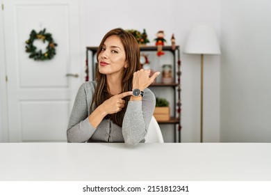 Young Latin Woman Sitting On The Table By Christmas Decor In Hurry Pointing To Watch Time, Impatience, Looking At The Camera With Relaxed Expression 