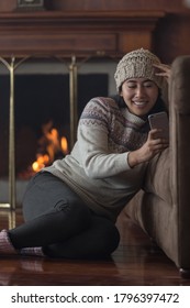 Young Latin Woman Smiling Sitting Sideways With A Cell Phone Holding Her Hand And Touching Her Head, Wears A Woolen Hat, Colored Jacket And Black Pants In The Background A Lit Fireplace