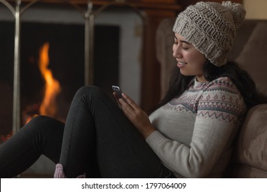 Young Latin Woman Smiling Sitting Sideways With A Cell Phone Holding Her Hand And Touching Her Head, With One Leg Stretched Out, Wears A Woolen Hat, Colored Jacket And Black Pants In The Background A 