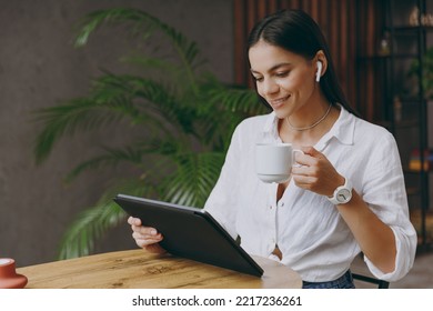 Young latin woman wear white shirt air pods listen music hold work on tablet pc computer drink coffee sit alone at table in coffee shop cafe restaurant indoors Freelance mobile office business concept