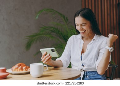 Young latin woman wear white shirt play app video games on mobile cell phone gadget smartphone do winner gesture sit at table in coffee shop cafe restaurant indoors. Freelance office business concept - Shutterstock ID 2217236283