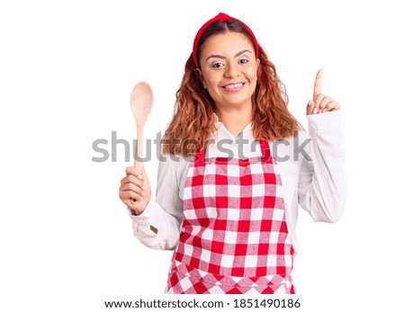 Young latin woman wearing apron holding wooden spoon surprised with an idea or question pointing finger with happy face, number one 