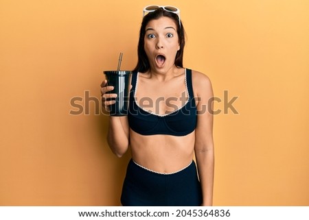 Young latin woman wearing bikini drinking soda scared and amazed with open mouth for surprise, disbelief face 
