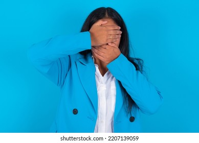 Young Latin Woman Wearing Blue Blazers Over Blue Background Covering Eyes And Mouth With Hands, Surprised And Shocked. Hiding Emotions.