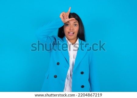Young latin woman wearing blue blazer over blue background gestures with finger on forehead makes loser gesture makes fun of people shows tongue