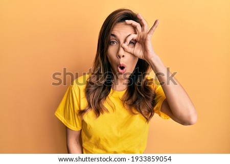 Young latin woman wearing casual clothes doing ok gesture shocked with surprised face, eye looking through fingers. unbelieving expression. 