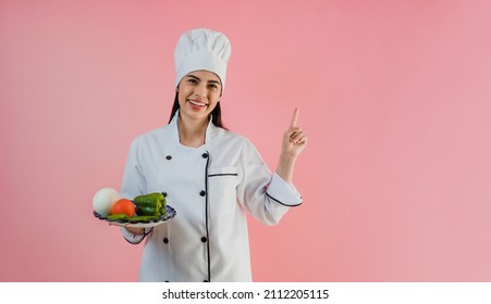 joven latinoamericana con sombrero de chef con ingredientes para cocinar comida mexicana con un fondo de color rosa