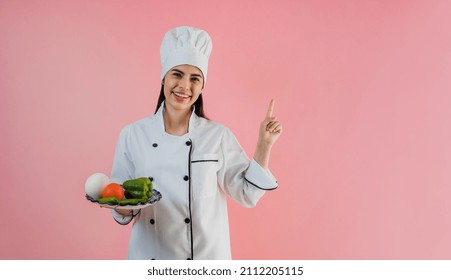 joven latinoamericana con sombrero de chef con ingredientes para cocinar comida mexicana con un fondo de color rosa