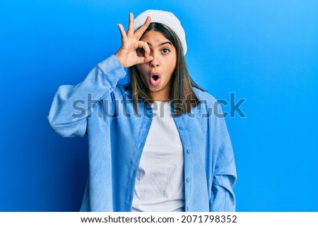 Young latin woman wearing cute wool cap doing ok gesture shocked with surprised face, eye looking through fingers. unbelieving expression. 