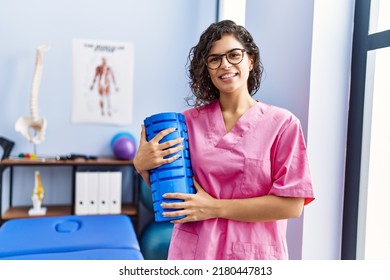 Young latin woman wearing physiotherapist uniform holding foam roller at clinic - Shutterstock ID 2180447813
