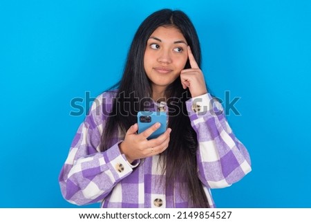 young latin woman wearing plaid shirt over blue background holding gadget while sticking out tongue