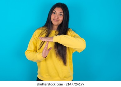 Young latin woman wearing yellow sweatshirt over blue background feels tired and bored, making a timeout gesture, needs to stop because of work stress, time concept.