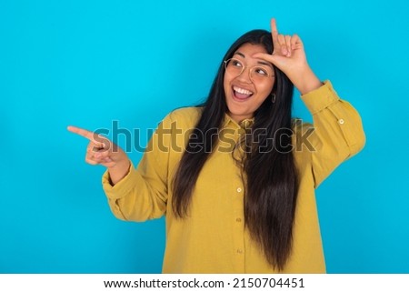 young latin woman wearing yellow shirt over blue background showing loser sign and pointing at empty space
