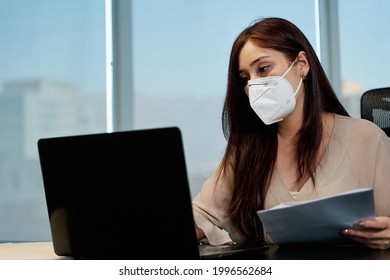 Young Latina Business Woman Wearing Covid-19 Protection Mask While Working On Her Laptop Inside The Office, Social Distancing And Work In Pandemic, Technology Concept