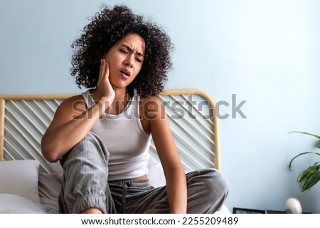 Young latina female with curly hair touching cheek in pain having toothache sitting on bed in cozy bedroom at home.