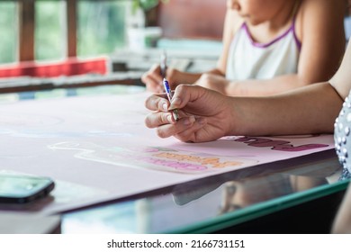 Young Latina Mother Teaching Her Daughter To Paint With Colored Pencils, Woman Doing School Homework With Her Young Daughter. Detailed View Of A Girl's Hand Holding A Pencil To Paint On The Poster.