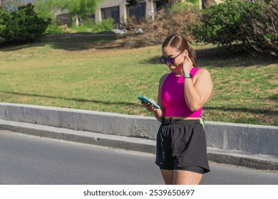 Young Latina woman adjusting earbuds while using smartphone outdoors in a park - Powered by Shutterstock