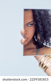 Young Latina woman with curly hair and a dress of straps of various colors posing in front of a mirror with the background a white wall. - Shutterstock ID 2259181815