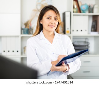 Young Latina Woman Doctor Assistant Standing In Medical Office Making Notes On Clipboard