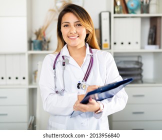 Young Latina Woman Doctor Assistant Standing In Medical Office Making Notes On Clipboard