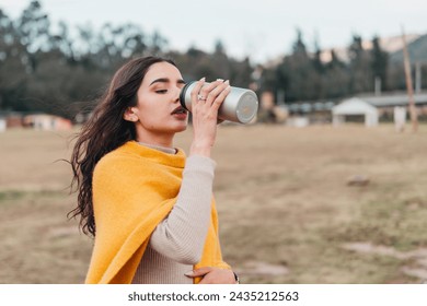 A young Latina woman sipping from a travel mug, wrapped in a cozy yellow sweater, outdoors with nature in the background - Powered by Shutterstock