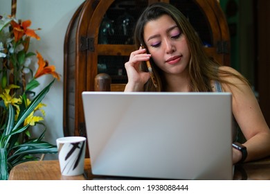 Young Latina Woman Using A Laptop And Making A Phone Call At Home While Drinking Coffee. Homeoffice Concept