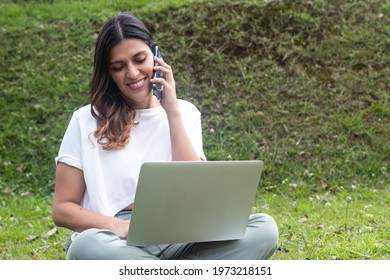 Young Latina Woman Using A Laptop And Making A Phone Call At The Park. New Normal Concept