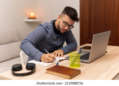 Young Latino Boy Writing His Homework In His Online College Class With His Computer And Book On The Table.