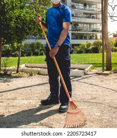 Young Latino Janitorial Worker In Blue Uniform Sweeping With Orange Rake In The Park 