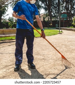 Young Latino Janitorial Worker In Blue Uniform Sweeping With Orange Rake In The Park 