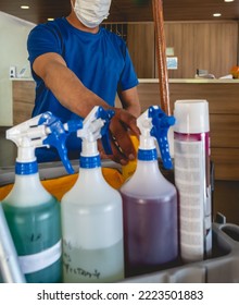 Young Latino Janitorial Worker In Blue Uniform And Trolley With Tools And Cleaning Supplies (sprayers, Broom, Cleaners, Etc)