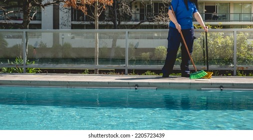 Young Latino Janitorial Worker Lady In Blue Uniform Sweeping And Cleaning At The Side Of Big Swimming Pool