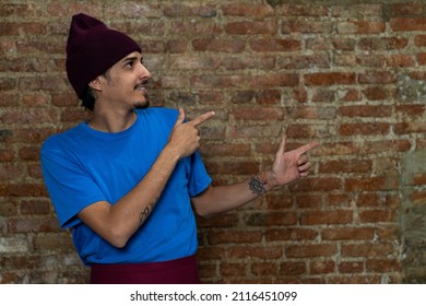 Young Latino Male (28) Looks And Points His Fingers To The Left Side. Copy Space, Brick Background. Concept Works