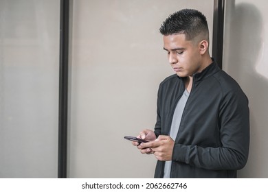 Young Latino Male Entrepreneur Looking At Social Networks On His Cell Phone Leaning Against A Glass Window, College Student Or Influencer With His Cell Phone In His Hand Shopping Online.