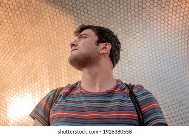 Young Latino Man With Beard, Short Hair And Shredded Shirt Looking To The Side And In The Background An Incredible Architectural Building With The Sun Reflecting On The Side.