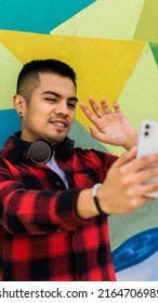 Young Latino Man Waving In Video Call With Cell Phone And Headphones, Colorful Background.