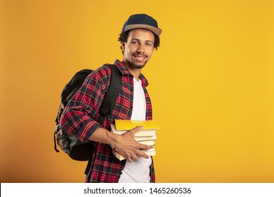 Young Latino Student Pose On Camera. Hold Books In Hand And Backpack. Handsome Positive Man Smile. Isolated Over Yellow Background