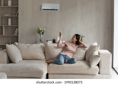 Young Latino Woman Relax On Sofa In Living Room Turn Electronic Conditioner With Remote Breathe Fresh Ventilated Air. Millennial Hispanic Female Renter Tenant Use Condition Device At Home Resting.