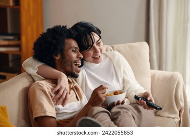 Young laughing man with bowl of popcorn and his wife with remote control sitting on couch in front of TV set and watching sitcom - Powered by Shutterstock