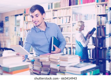 Young Laughing Man Choosing New Book From Many In Book Shop