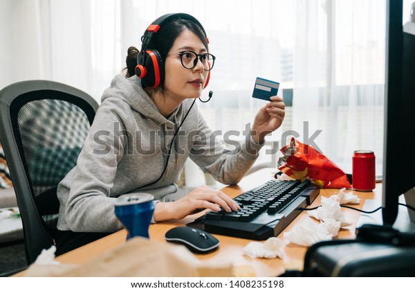 young lazy college girl nerd at home on summer\
break off from school holding credit card paying online e commerce\
buying game points. asian woman geek with headphones playing\
computer internet.