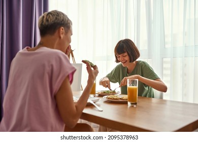 Young lesbian caucasian couple having breakfast at wooden table at home. Concept of domestic lifestyle. Idea of homosexual relationship and spending time together. Modern apartment. Sunny morning time - Powered by Shutterstock