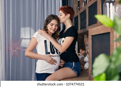 Young lesbian couple expecting a baby. A pretty woman hugging and kissing her pregnant girlfriend. The mom-to-be smiling, her hands resting on the belly. Future same-sex parents enjoying togetherness.