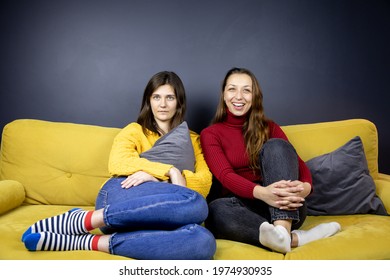 Young Lesbian Couple Sit On Comfy Sofa Knees Bent Smiling Watching TV Huddled Together. LGBT Family Resting Lazy Evening At Home On Couch. Same Sex Romantic Date Concept Copy Space