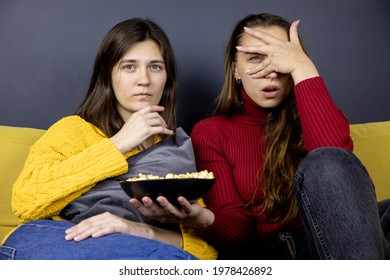 Young Lesbian Couple Sit On Comfy Sofa Knees Bent Huddled Together Frightened Watching TV Peeking Covering Eyes With Palm. LGBT Family Resting Lazy Evening At Home On Couch. Same Sex Romantic Date