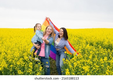 A Young Lesbian LGBT Couple With A Child Smiles While Holding A Rainbow Flag Of Pride
