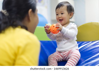 Young Little Asian Baby Holding Balls And Toys Playing With Female Preschool Teacher In Classroom. Kid Looking At Nanny That Teaching Her With Smile, Laughing And Enjoy Participating In The Class.