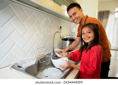 Young little asian daughter helping her father in the kitchen washing dishes and looking at camera with happy, after breakfasting during Ramadan at home. Parenthood, healthy lifestyle and islamic - Powered by Shutterstock