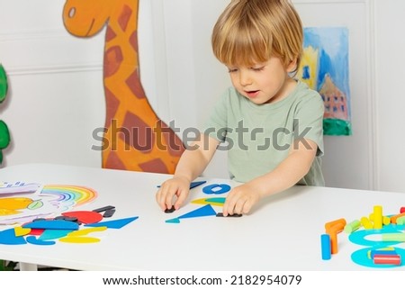 Young little blond boy form shapes standing by the table in the kindergarten playing developmental games